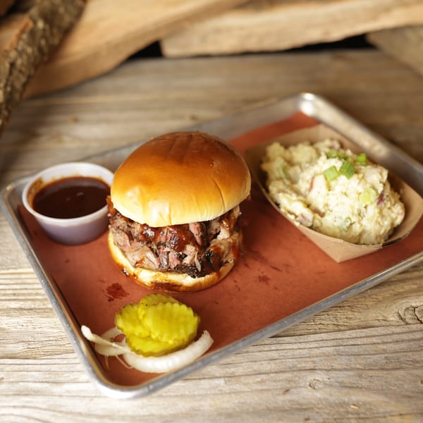 Cedar River BBQ pulled pork sandwich with potato salad, pickles, onions, and BBQ sauce on a tray.